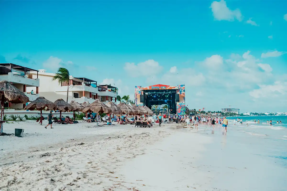 Wide view of beach with resort, palapas, and concert stage in the distance during daytime.