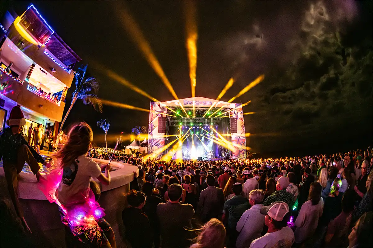 Crowd watching nighttime beach stage concert with bright lights shining over the audience.