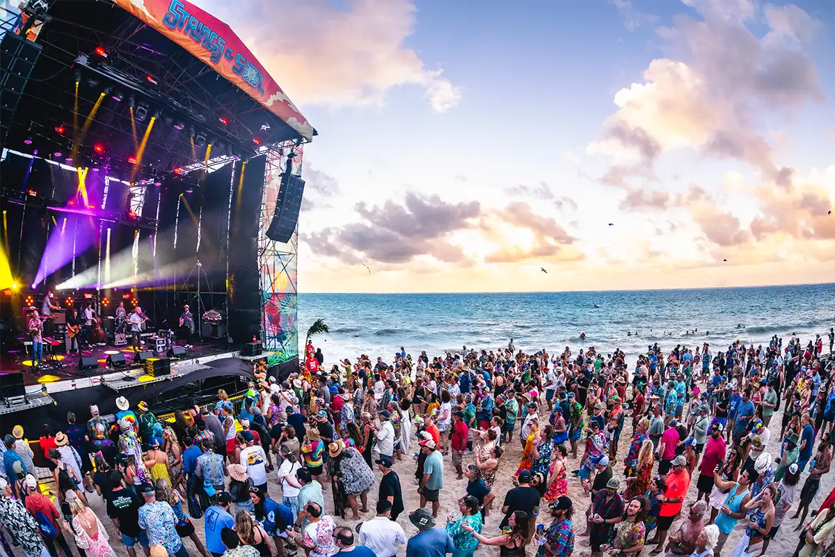 Crowd gathered on the beach watching concert at sunset by the ocean.