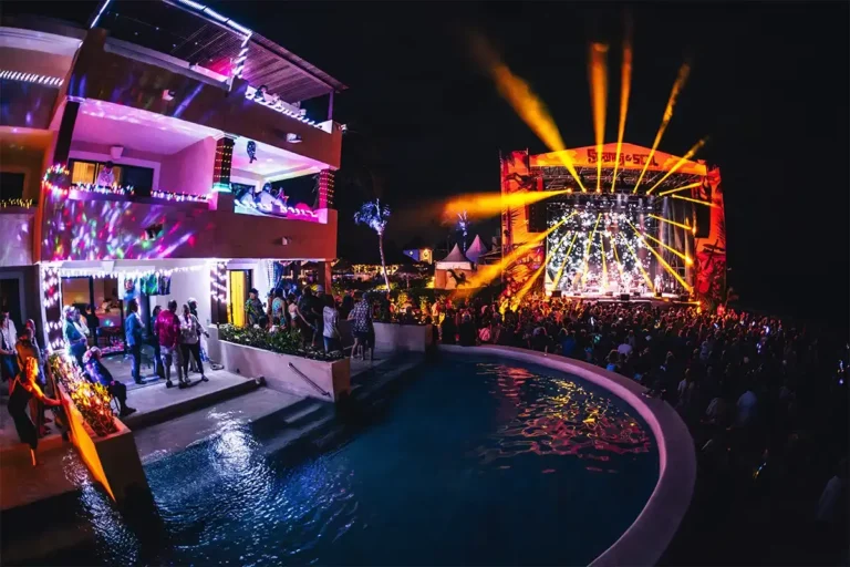 Nighttime concert at beach stage with crowd, pool, and resort building in the foreground