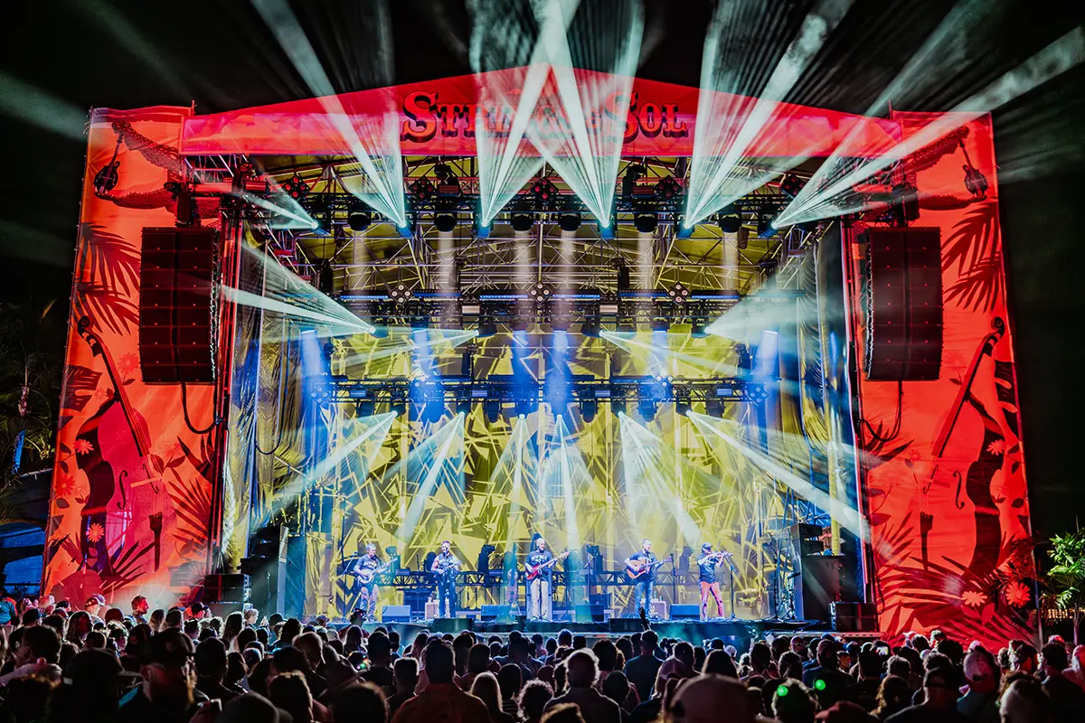 Nighttime beach stage concert with bright lights shining into the sky.
